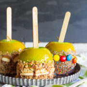 Three caramel apples with nuts and candy toppings, displayed on a metal tray with a dark background.