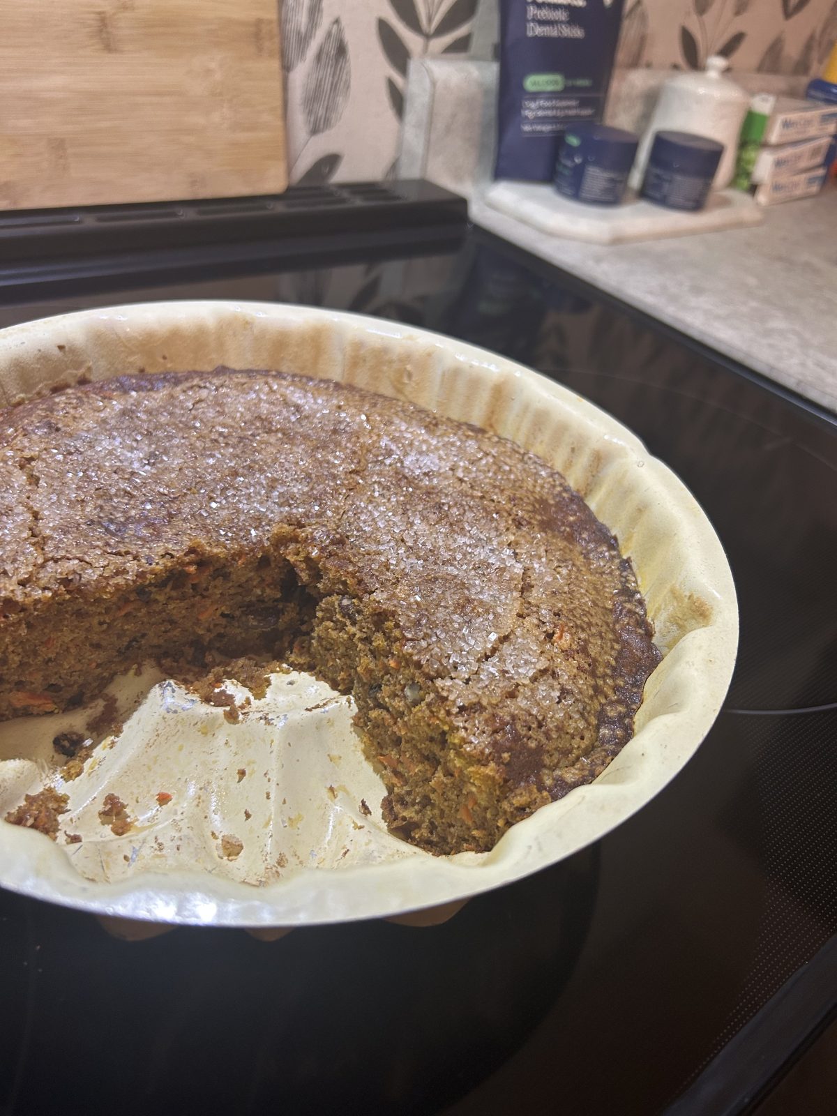 A round cake with a slice missing sits in a white baking dish on a stovetop. The cake has a sugary crust and visible carrot bits. Kitchen items are in the background.