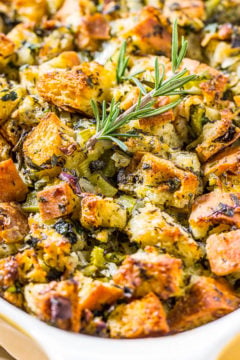 A baking dish filled with golden-brown cubed stuffing garnished with fresh rosemary.