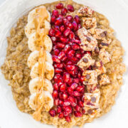A bowl of oatmeal topped with banana slices, pomegranate seeds, and a drizzle of peanut butter.