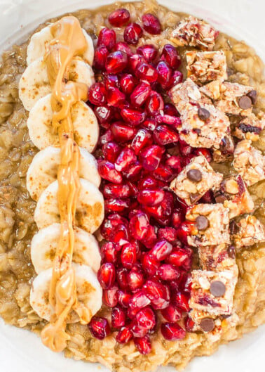 A bowl of oatmeal topped with banana slices, pomegranate seeds, and a drizzle of peanut butter.