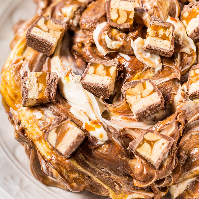 A close-up of a decadent dessert featuring layers of pastry topped with caramel, nuts, and chunks of chocolate bars.