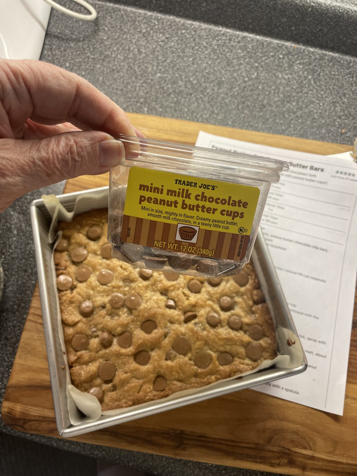 Hand holding a container of Trader Joe's mini milk chocolate peanut butter cups above a baked square dessert in a pan on a wooden board.