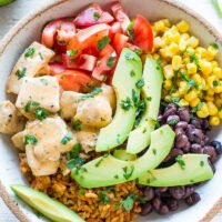 A bowl containing a colorful mix of rice, beans, corn, tomatoes, avocado, and diced chicken, garnished with herbs.
