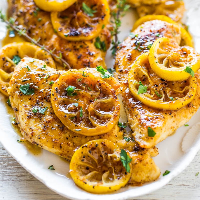 Grilled chicken breasts topped with lemon slices and herbs on a white plate.