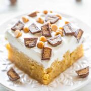 A slice of frosted cake topped with chocolate pieces and caramel bites on a white plate.