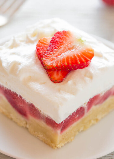 A slice of strawberry shortcake topped with whipped cream and a strawberry slice on a white plate.