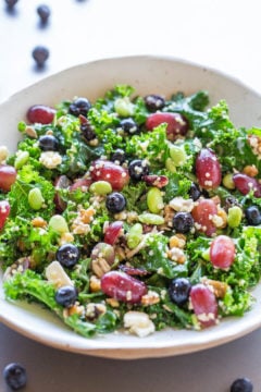 A colorful kale salad with beans, blueberries, edamame, and nuts in a white bowl.