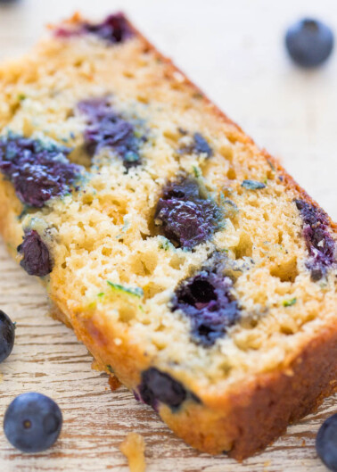 A slice of blueberry loaf cake with whole blueberries scattered around.