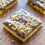 A close-up of a square chocolate and nut topped shortbread dessert on a wooden surface.