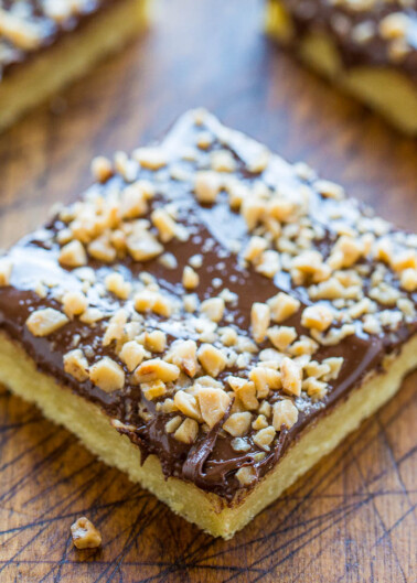 A close-up of a square chocolate and nut topped shortbread dessert on a wooden surface.