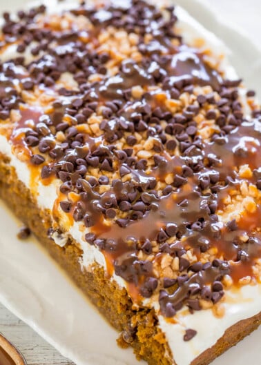 A rectangular carrot cake topped with cream cheese frosting, caramel drizzle, chopped nuts, and mini chocolate chips.