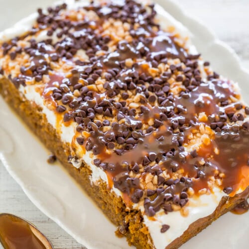 A rectangular carrot cake topped with cream cheese frosting, caramel drizzle, chopped nuts, and mini chocolate chips.