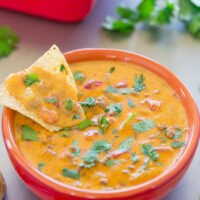 A bowl of creamy, chunky dip garnished with cilantro, with a tortilla chip dipped inside, accompanied by more chips in the background.