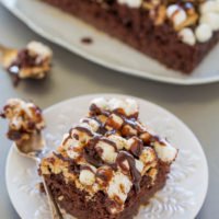 A slice of chocolate cake topped with marshmallows, nuts, and drizzled with chocolate syrup, served on a white plate with a fork, with more cake in the background.