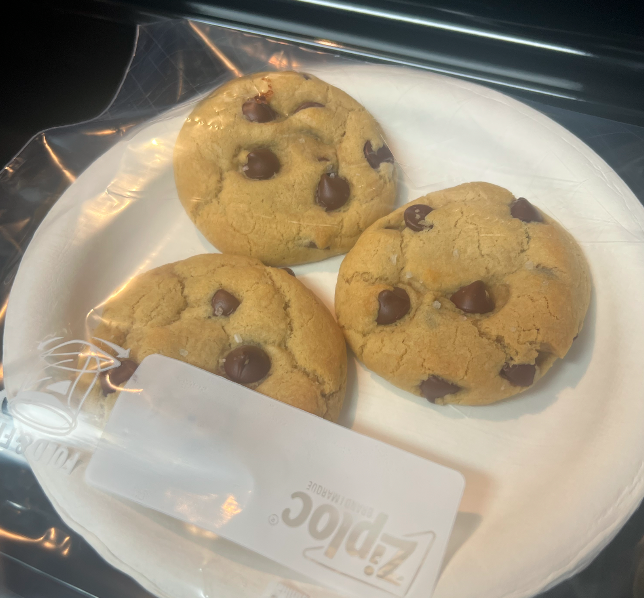 Three chocolate chip cookies on a white paper plate, partially covered by a clear plastic Ziploc bag.