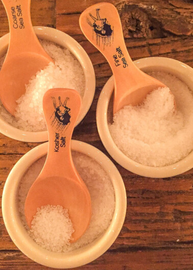 Three small bowls of salt with wooden spoons on a wooden table.