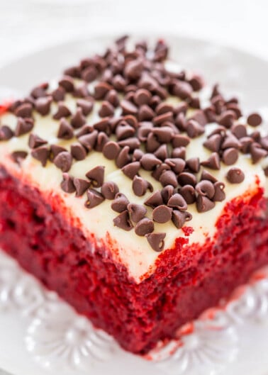 A slice of red velvet cake with cream cheese frosting and chocolate chips on top, served on a white plate.