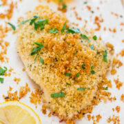 Breaded chicken breast garnished with parsley and lemon on a white background.