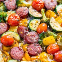 A colorful mix of roasted vegetables and sliced sausage topped with grated cheese on a baking tray.