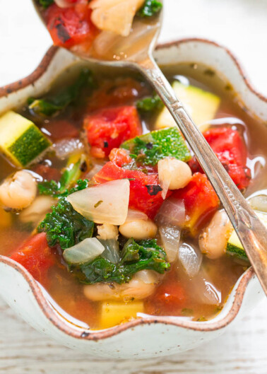 A colorful bowl of vegetable soup with tomatoes, zucchini, kale, onions, and beans, served with a spoon.