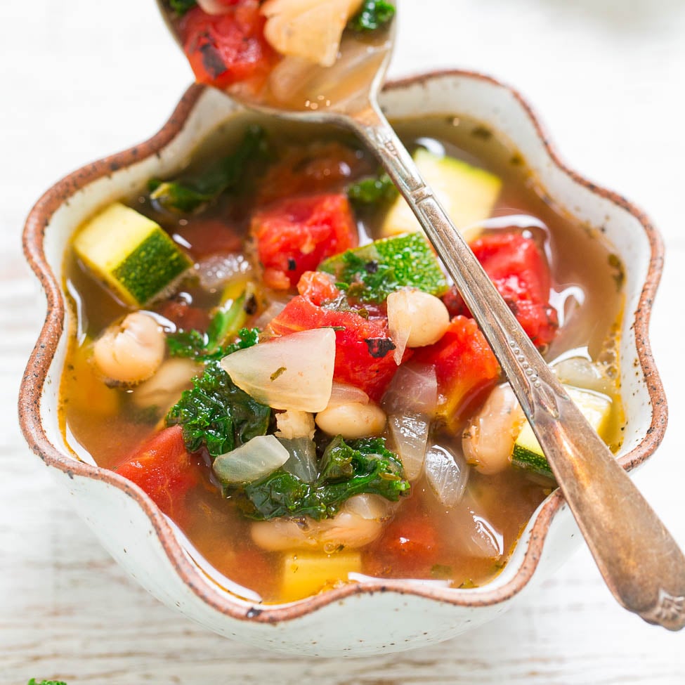 A colorful bowl of vegetable soup with tomatoes, zucchini, kale, onions, and beans, served with a spoon.