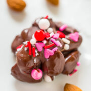Chocolate-covered confectionery adorned with red and pink sprinkles, hearts, and white balls on a white background with almonds nearby.