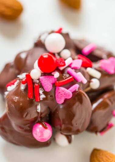 Chocolate-covered confectionery adorned with red and pink sprinkles, hearts, and white balls on a white background with almonds nearby.