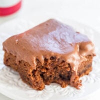 A piece of chocolate frosted cake on a white, ornate plate with a bite taken out of it, and a red can in the background.