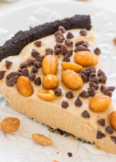 A slice of peanut butter pie topped with chocolate chips and peanuts on an oreo crust, served on a white plate.