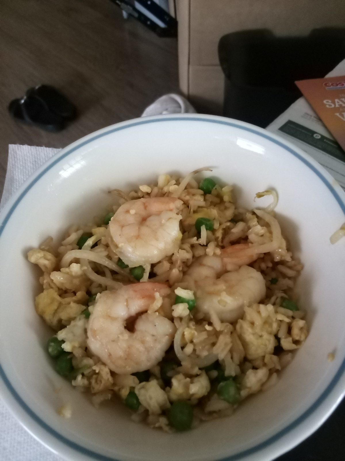 A bowl of shrimp fried rice with peas, scrambled eggs, and bean sprouts, placed on a table with various items in the background.