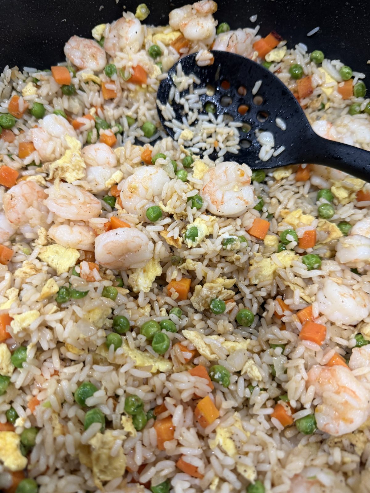 Shrimp fried rice with peas, carrots, scrambled egg, and a black slotted spoon.