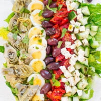 A colorful mediterranean salad with artichokes, eggs, olives, roasted peppers, cucumbers, and feta cheese, neatly arranged on a white surface.