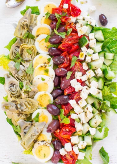 A colorful mediterranean salad with artichokes, eggs, olives, roasted peppers, cucumbers, and feta cheese, neatly arranged on a white surface.