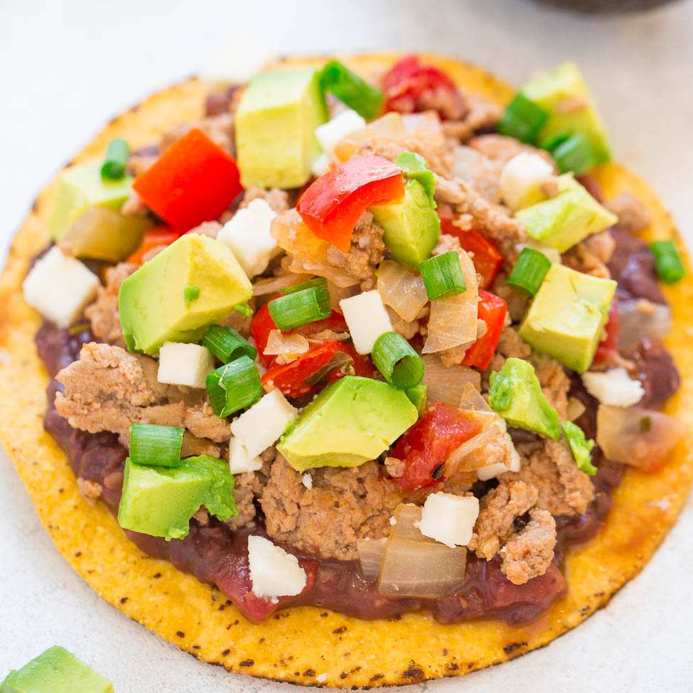 A tostada topped with beans, ground meat, avocado, tomatoes, and diced onions.