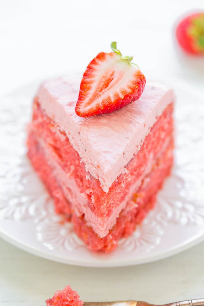 A slice of strawberry cake topped with a half strawberry on a white plate.