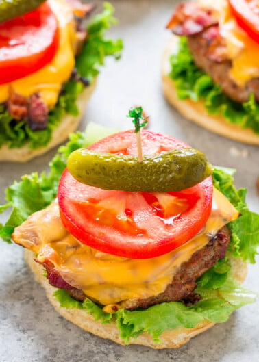 Two cheeseburgers with lettuce, tomato, pickles, and bacon, garnished with a gherkin on top.