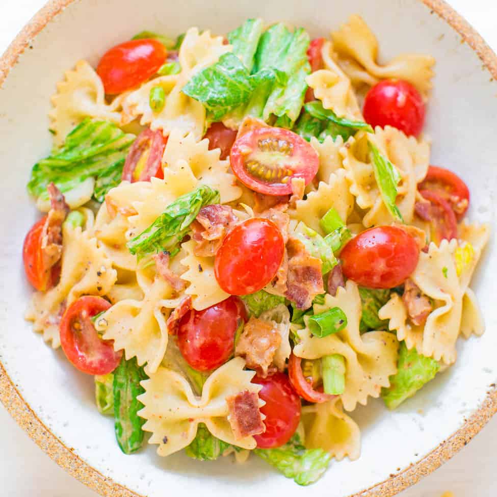 A bowl of bow-tie pasta salad with cherry tomatoes, greens, and bacon bits.