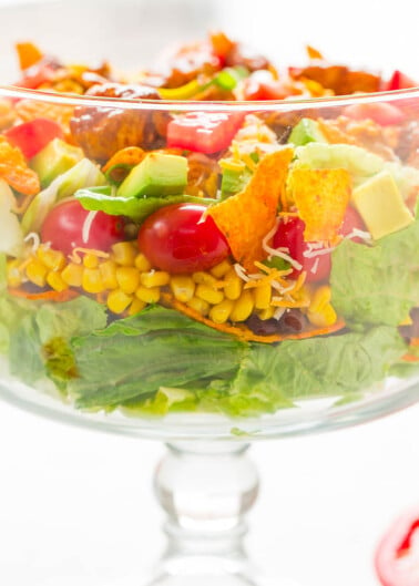 A colorful taco salad with lettuce, tomatoes, corn, avocado, shredded cheese, and seasoned meat in a glass bowl.