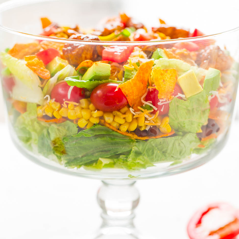 A colorful taco salad with lettuce, tomatoes, corn, avocado, shredded cheese, and seasoned meat in a glass bowl.