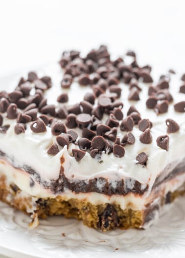 A slice of chocolate chip cookie cake topped with white frosting and mini chocolate chips on a white plate.