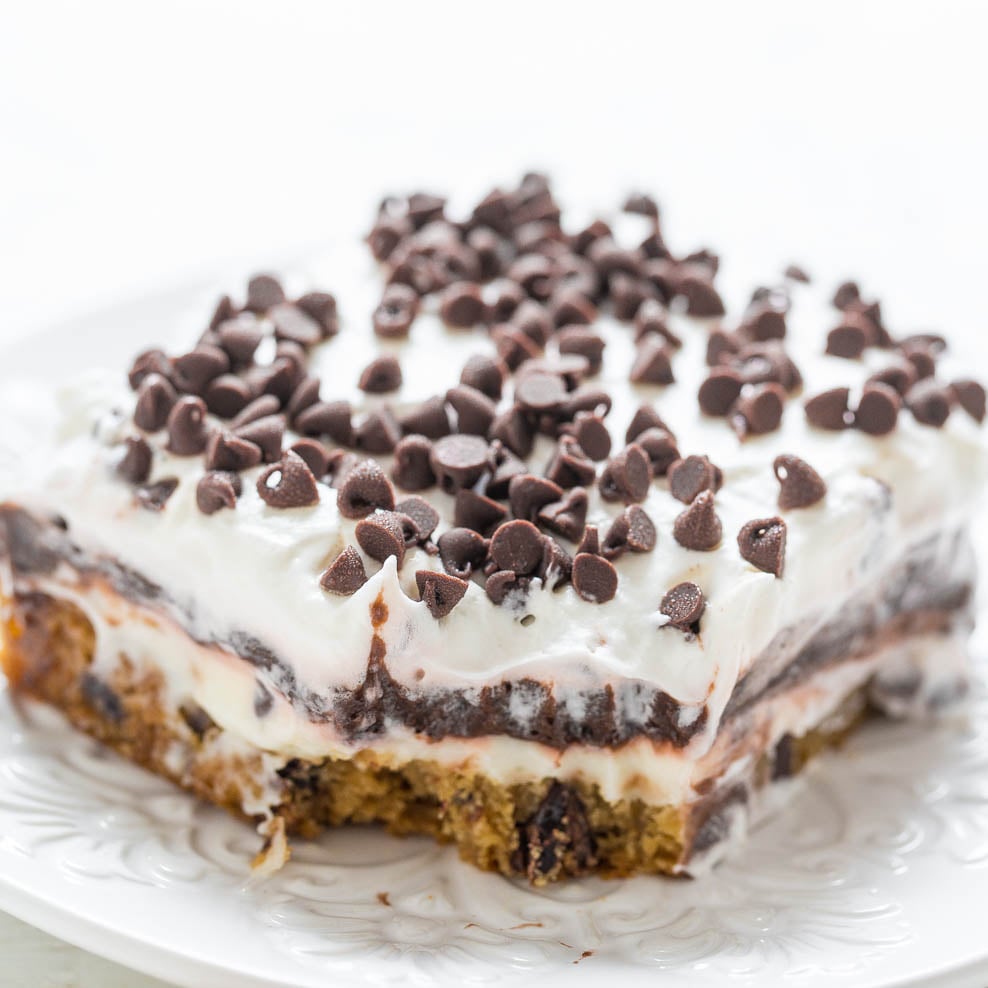 A slice of chocolate chip cookie cake topped with white frosting and mini chocolate chips on a white plate.