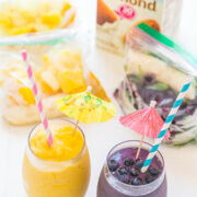 Two smoothies with paper umbrellas and striped straws, one yellow and one purple, with ingredients in the background.