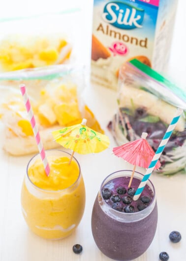Two smoothies with paper umbrellas and striped straws, one yellow and one purple, with ingredients in the background.