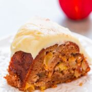 A slice of apple cake with cream cheese frosting on a white plate, with an apple in the background.