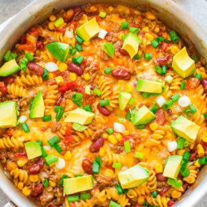 A skillet filled with a colorful pasta dish featuring rotini, beans, corn, diced tomatoes, melted cheese, and topped with avocado cubes and chopped green onions. An avocado half is seen beside the skillet.