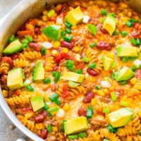 A skillet filled with cheesy taco pasta topped with diced avocado and sliced green onions.