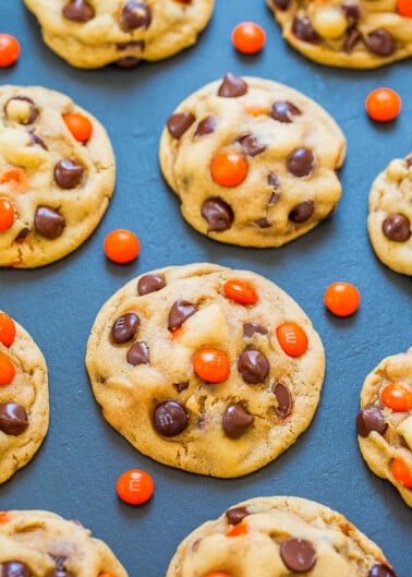 Freshly baked chocolate chip cookies with orange-colored candies on a dark surface.