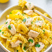 A bowl of chicken pasta garnished with parsley, with a lemon and herbs in the background.