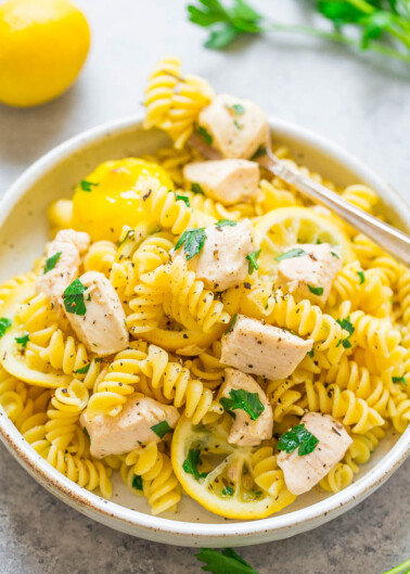 A bowl of chicken pasta garnished with parsley, with a lemon and herbs in the background.